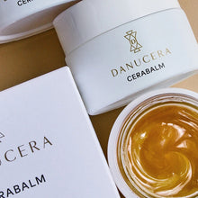 Load image into Gallery viewer, Danucera Cerabalm skincare products including a white jar with gold lettering, a box, and an open jar showing the golden-yellow balm inside, arranged on a beige surface.
