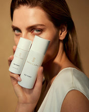 Load image into Gallery viewer, A woman holds two white Danucera D22 Tonic bottles in front of her face against a warm beige background.

