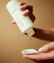 Load image into Gallery viewer, A close-up of hands holding a white bottle of Danucera D22 Tonic, with a single drop of clear liquid dripping onto a white handkerchief against a warm beige background.
