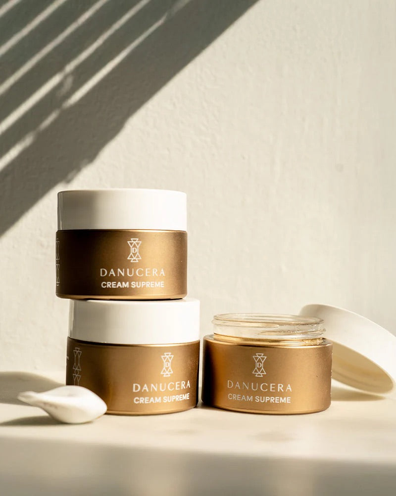 Three Danucera Cream Supreme jars in a stacked and open arrangement on a warm beige surface with soft shadows and a white lid resting nearby.