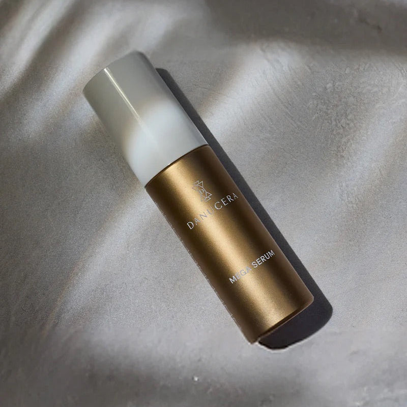 A bottle of Danucera's Magic Serum with a metallic gold body and a frosted white cap, resting on a textured gray surface with soft shadows.
